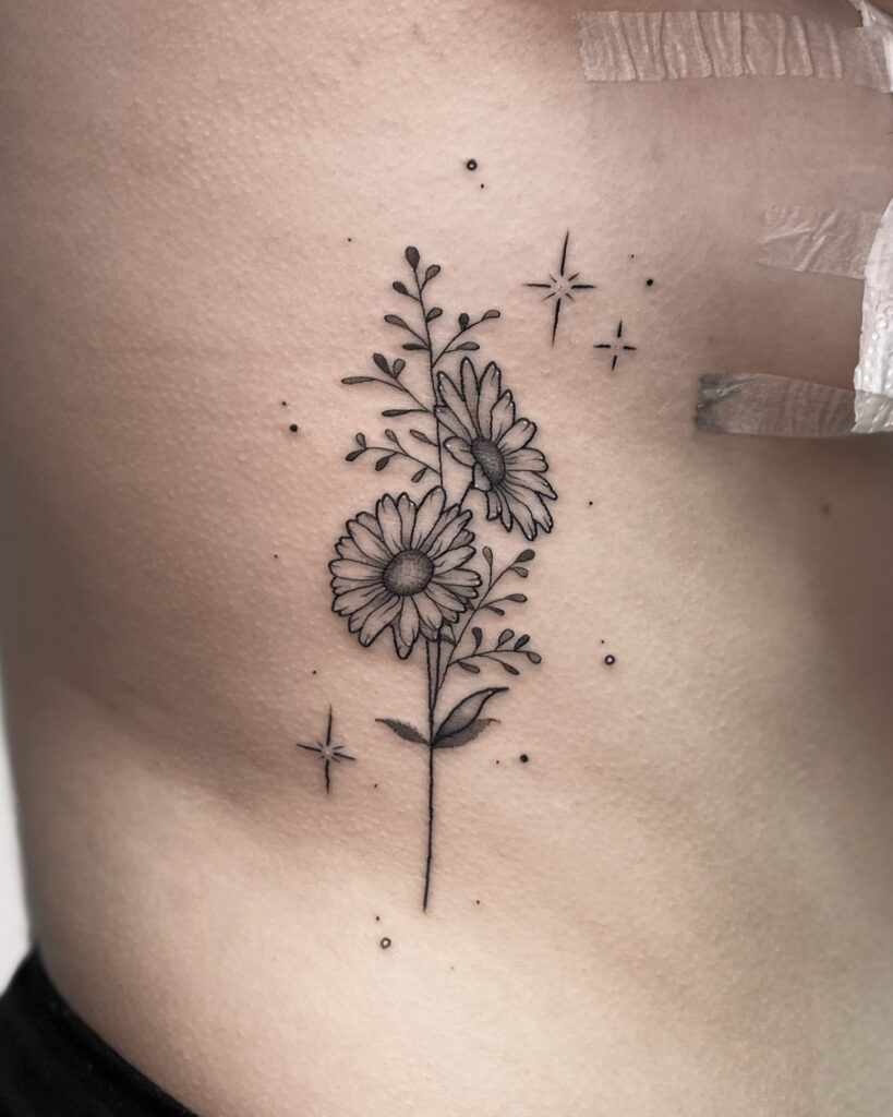 This delicate floral tattoo features two beautifully detailed daisy flowers with fine linework and subtle dot shading. The design in enhanced with tiny sparkles and dots, adding an ethereal touch. Placed on the ribcage, this minimalist black ink tattoo in perfect choice for those who love elegant and feminine designs. Floral tattoos symbolize growth, beauty and new beginnings, making this piece both aesthetic and meaningful.