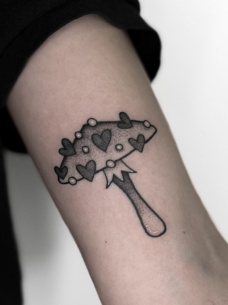 A detailed black ink tattoo of mushroom adorned with heart shapes and pearl-like accents, located on the outer forearm. This design blends fantasy and nature-inspired elements, creating a unique and artistic look. Perfect for those who love whimsical yet edgy tattoo styles.