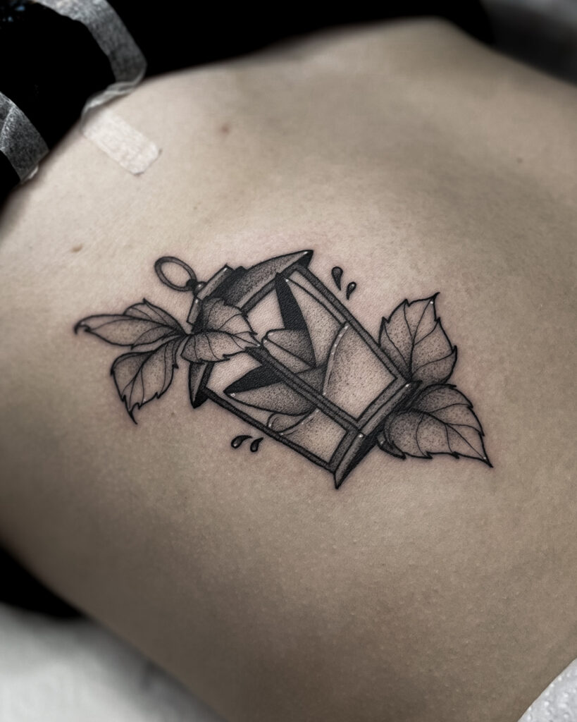 Black and grey lantern tattoo and little paper boat featuring fine linework, bold shading and botanical elements. This detailed lantern design is perfect for nature-inspored tattoos. The placement on the upper back makes it ideal for medium sized tattoo ideas. Clean lines and smooth dootwork are suitable for both men and women. Symolizing guidance, light and protection